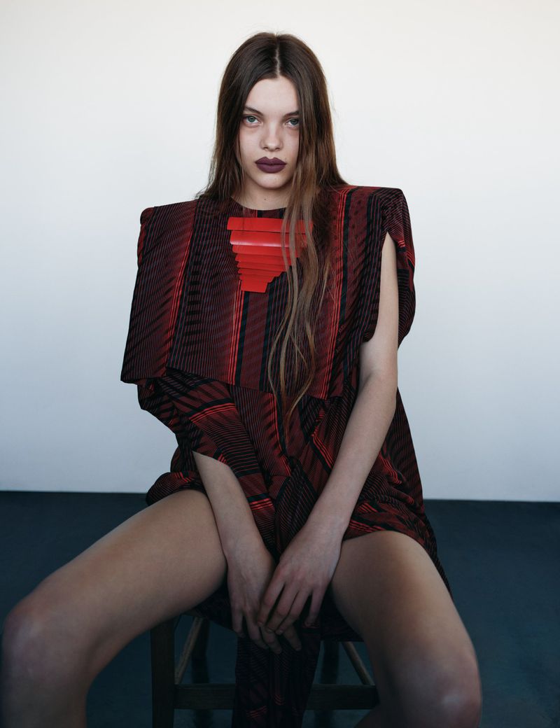 Fashion portrait of a woman with long brown hair, dark lipstick, and a red geometric dress, sitting on a wooden stool in front of a white wall.