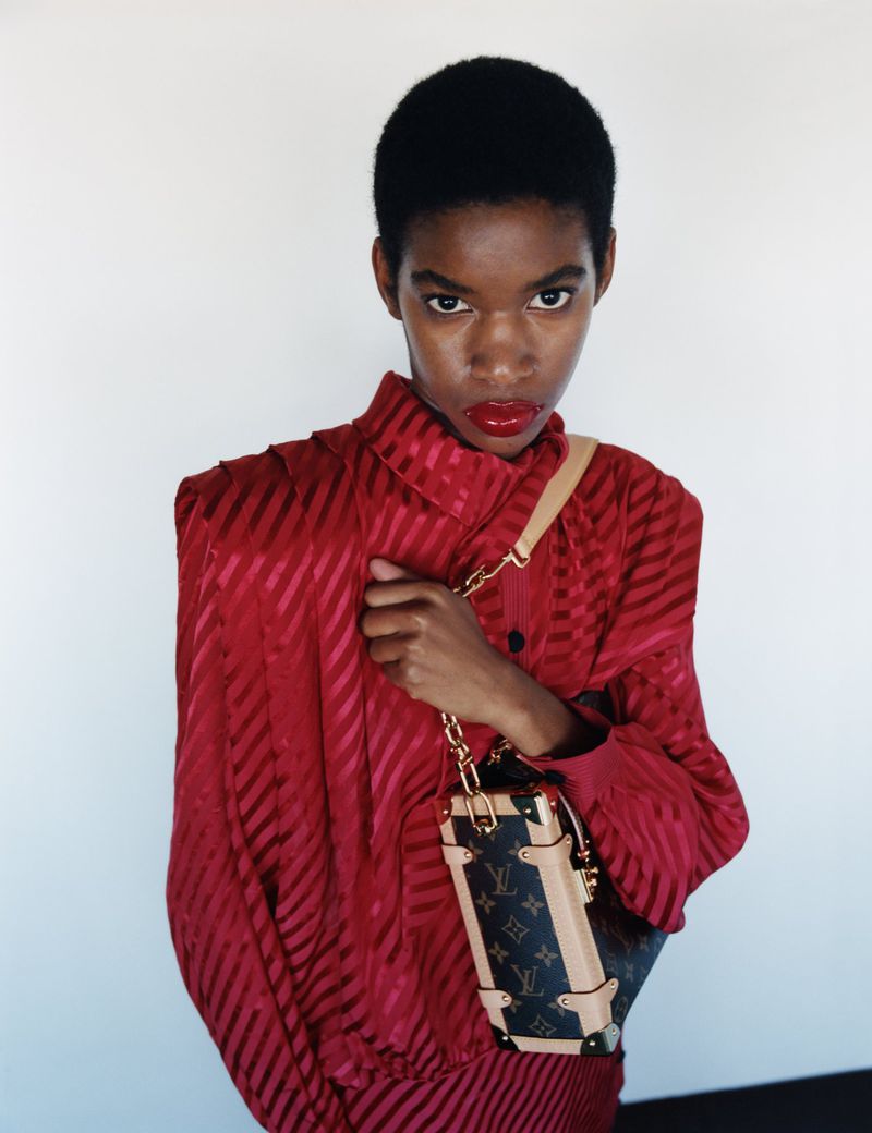 An African-American woman in a red striped dress and matching red lipstick, posing with a Louis Vuitton Petite Malle Souple handbag in a studio.