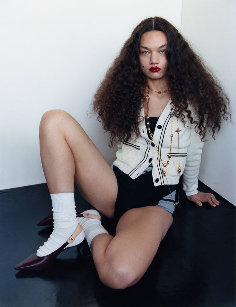 A curly-haired woman in a cardigan, black shorts, and burgundy shoes poses indoors against a white wall and black floor.