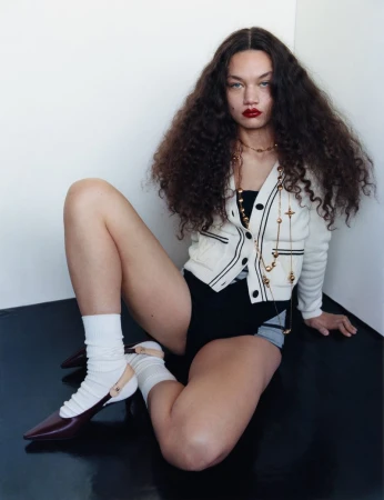 A curly-haired woman in a cardigan, black shorts, and burgundy shoes poses indoors against a white wall and black floor.
