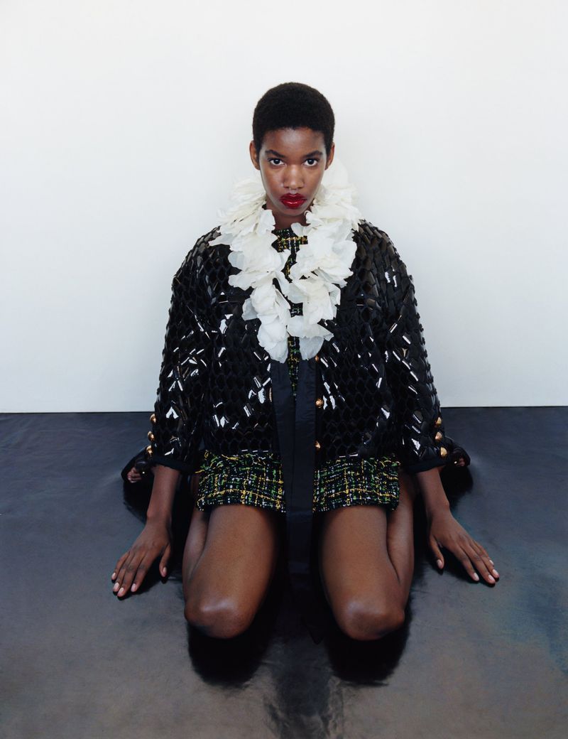 A black female model in a stylish black jacket and green tweed shorts poses in a studio with dark red lipstick and a white collar.