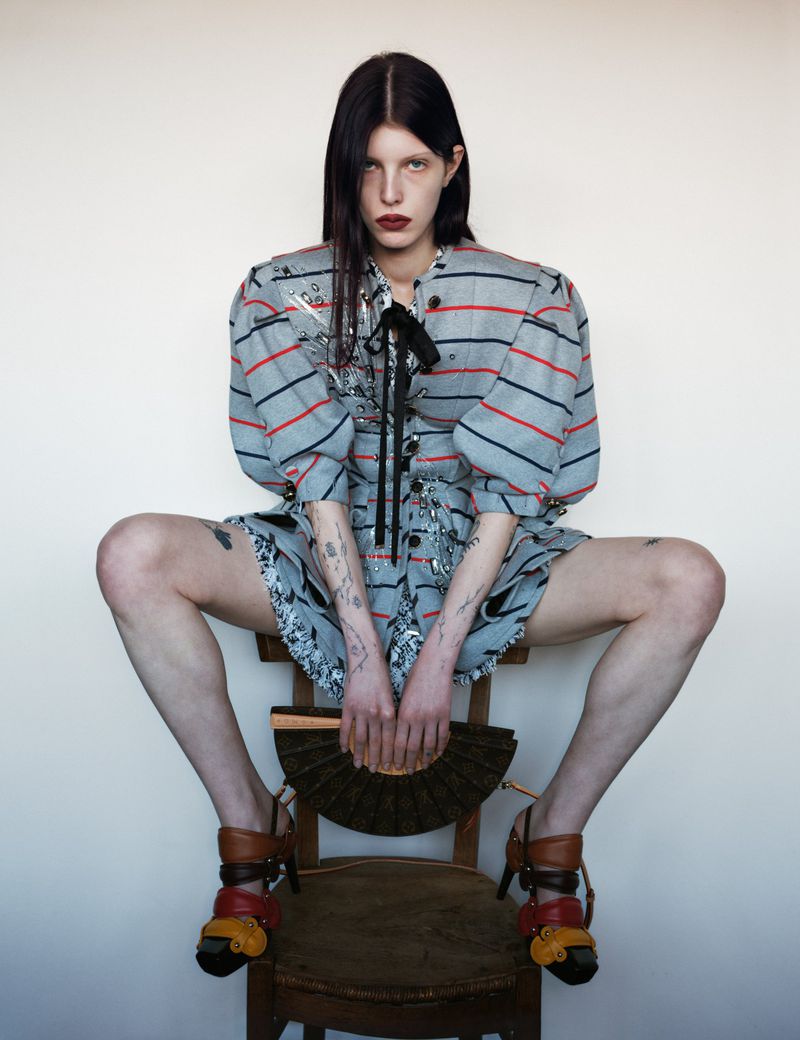 A slender woman with dark hair poses on a wooden chair, wearing a patterned dress with bold shoes and holding a folding fan.
