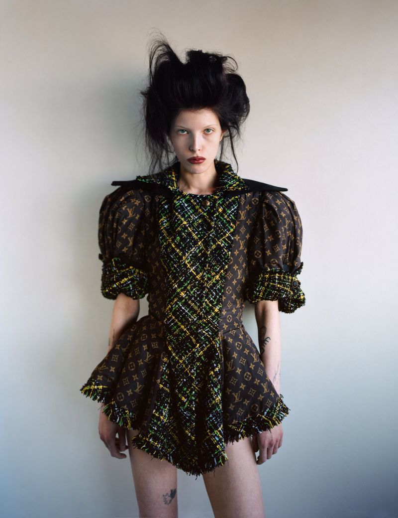 Woman in elaborate brown patterned dress with puffy sleeves and dark hair, standing indoors against a neutral-toned wall.