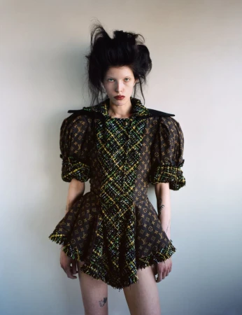 Woman in elaborate brown patterned dress with puffy sleeves and dark hair, standing indoors against a neutral-toned wall.
