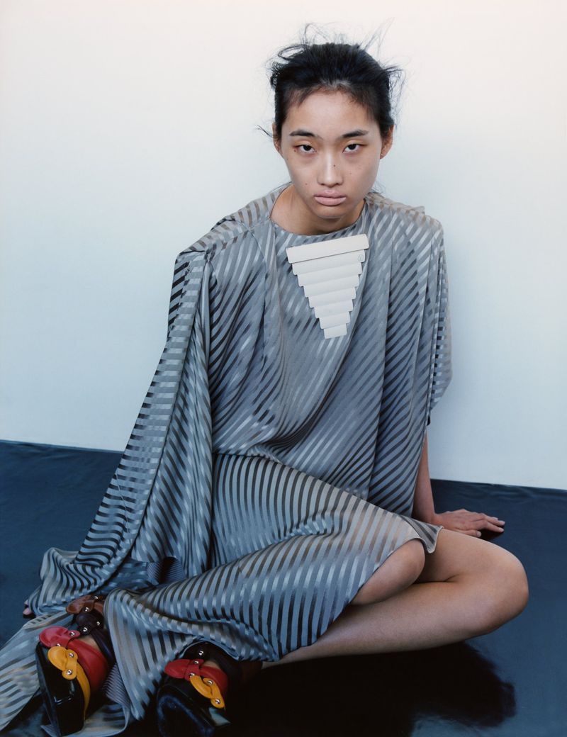 A seated Asian woman in a grey striped dress and unique platform shoes poses against a white background in a fashion editorial photograph.