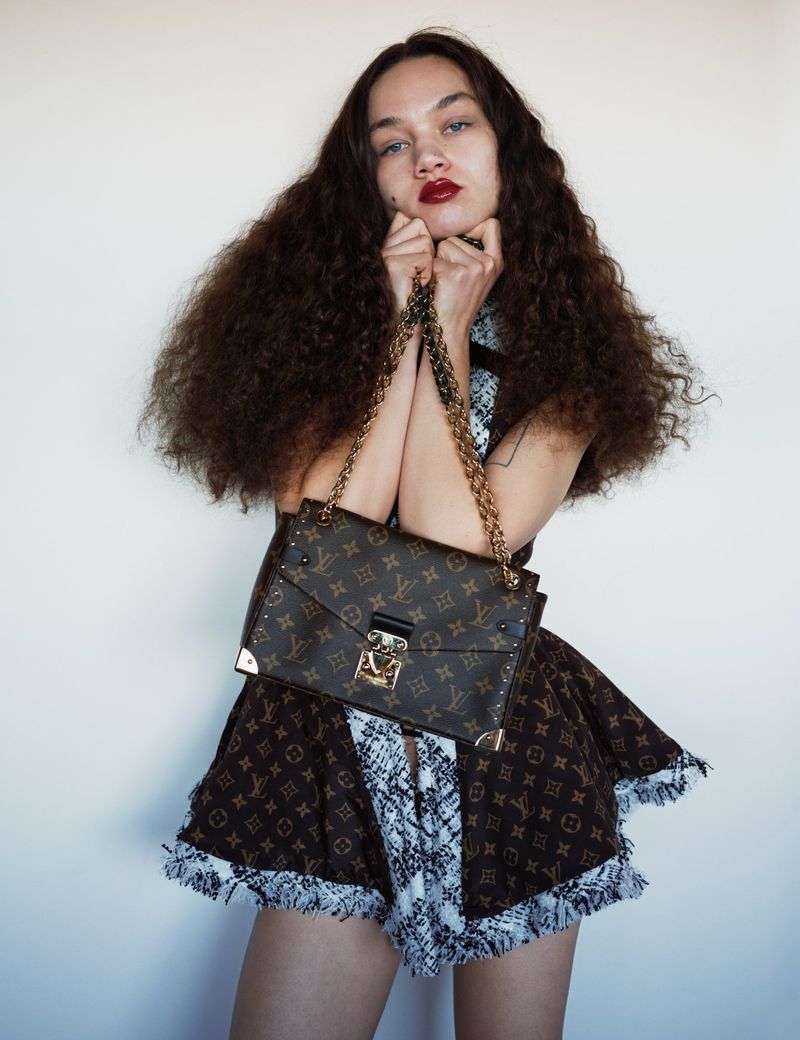 Brunette woman in a brown dress and a designer purse poses against a white wall in a studio with hands near her face.