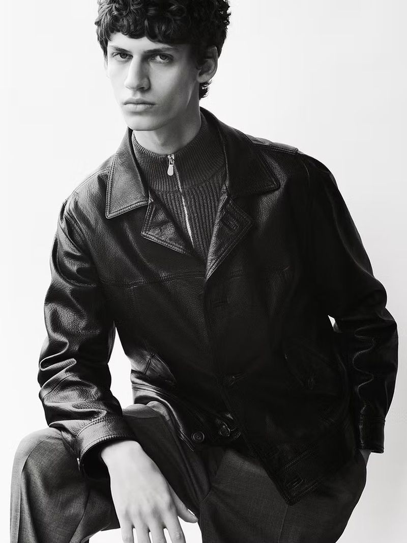 Monochrome studio shot of a male model with curly hair, wearing a leather jacket and turtleneck, posing against a white background.