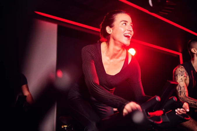 Woman smiles while exercising on a stationary bike in a dimly lit gym with red lighting, surrounded by other fitness enthusiasts.