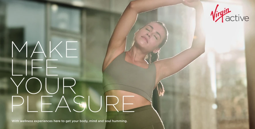 Woman in a green sports bra stretching her arms overhead in a bright sunlit fitness space for Virgin Active