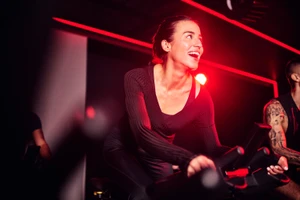 Smiling woman in dark athletic wear riding a stationary exercise bike in a dark gym illuminated by red lighting.