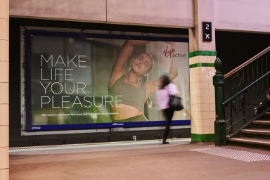 Large outdoor advertising billboard for Virgin Active reading Make Life Your Pleasure displayed at a transport station platform.