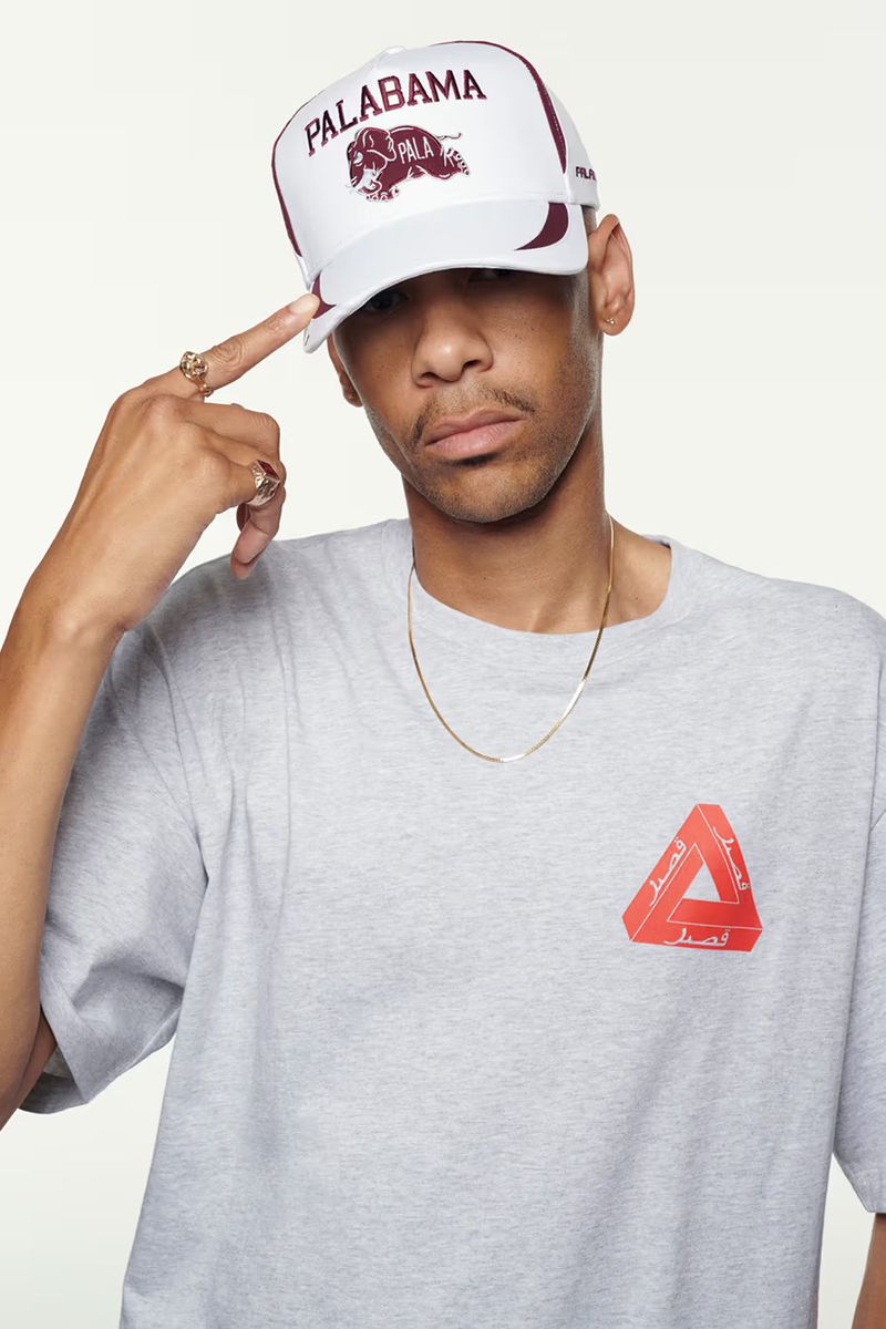 Adult male in a grey t-shirt and baseball cap touches his head in a studio shot with a white background, wearing a gold necklace.