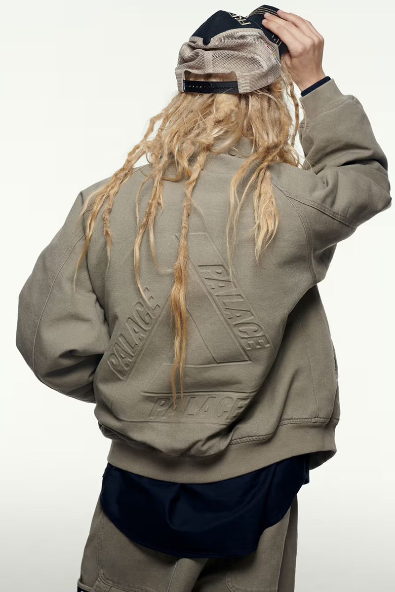 Back view of a person with blonde dreadlocks wearing a baseball cap, grey jacket with embossed logo and shorts against a white background.