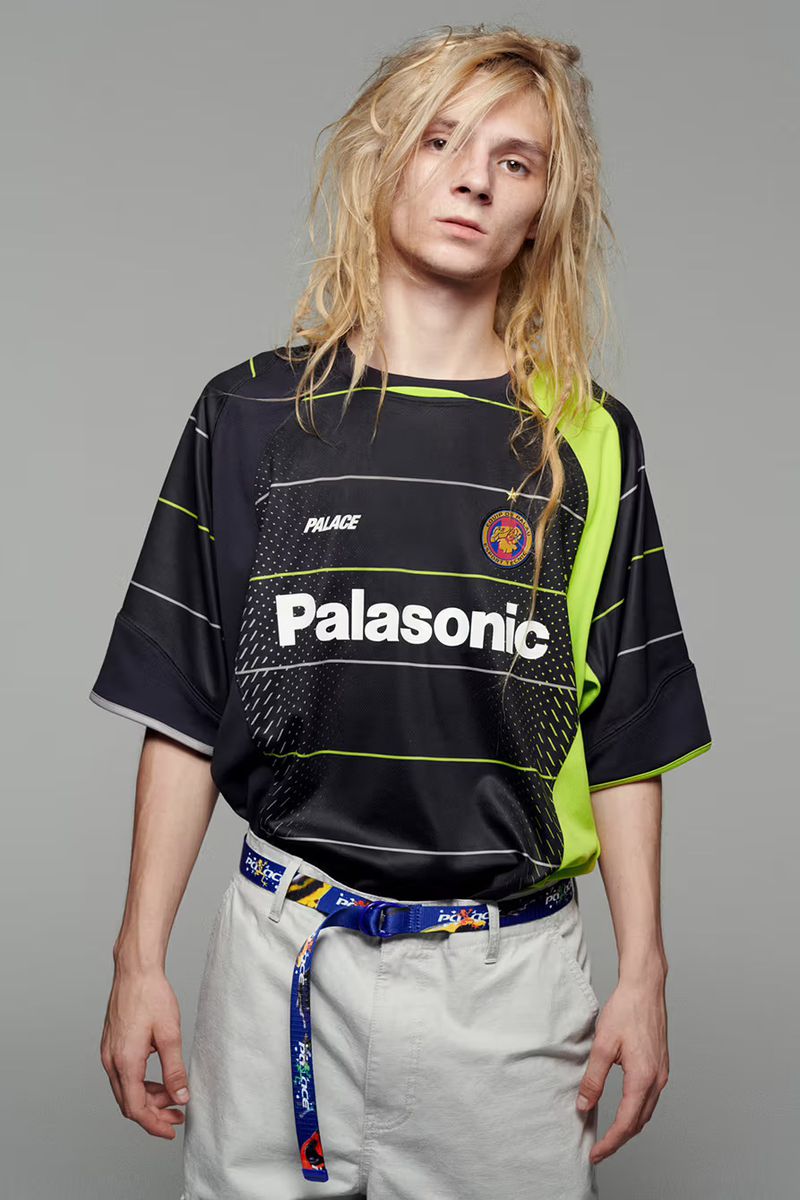 Male fashion model with long blonde hair poses in a black Palasonic soccer jersey with a patterned belt against a grey backdrop.