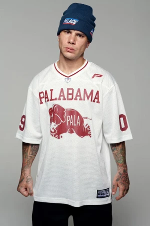 A man in a blue beanie and a white jersey with a graphic of an elephant stands indoors against a neutral background.