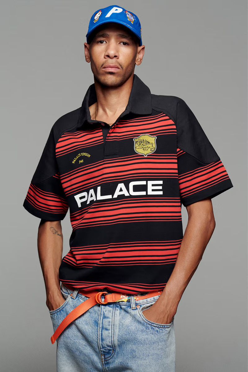 Adult male posing in a black and red striped polo shirt, light blue jeans, blue baseball cap, and orange belt against a grey backdrop.