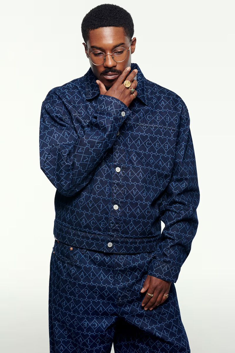 Portrait of a Black man in a patterned denim jacket and pants, wearing gold glasses and rings, against a plain white background.