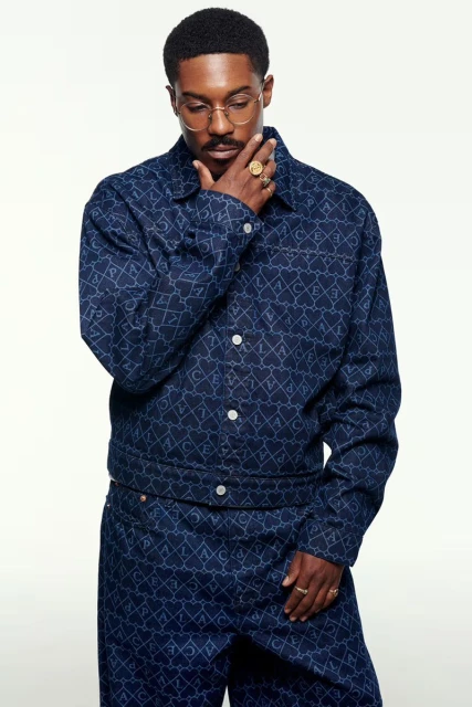 Portrait of a Black man in a patterned denim jacket and pants, wearing gold glasses and rings, against a plain white background.