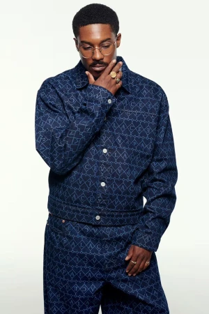 Portrait of a Black man in a patterned denim jacket and pants, wearing gold glasses and rings, against a plain white background.