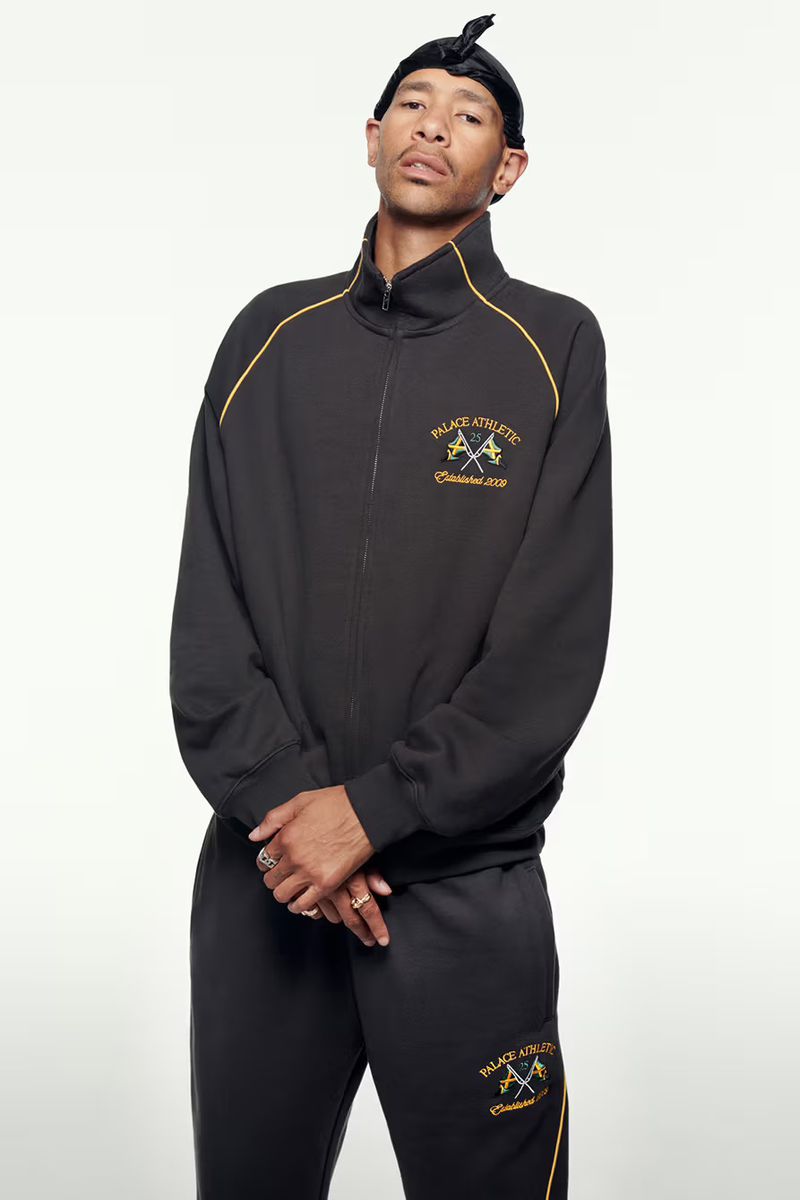 Adult male in a black athletic tracksuit with gold accents and a black durag poses in a studio with a neutral background.