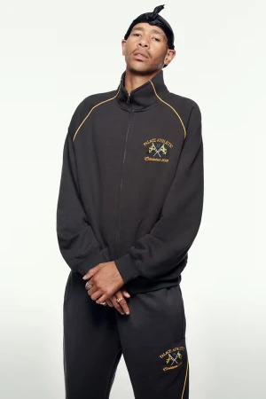 Adult male in a black athletic tracksuit with gold accents and a black durag poses in a studio with a neutral background.