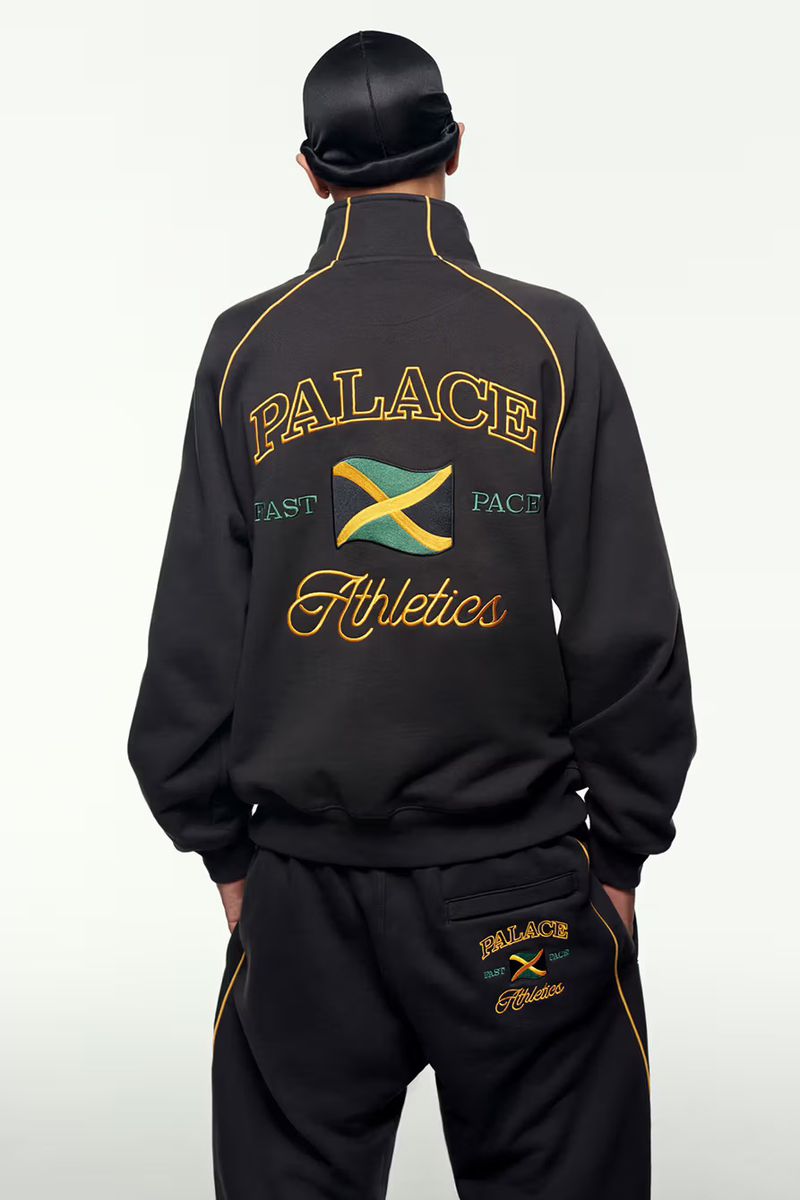 Person wearing a black Palace tracksuit featuring gold, green, and yellow detailing and a Jamaican flag emblem against a white backdrop.