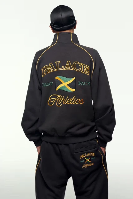 Person wearing a black Palace tracksuit featuring gold, green, and yellow detailing and a Jamaican flag emblem against a white backdrop.