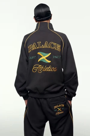 Person wearing a black Palace tracksuit featuring gold, green, and yellow detailing and a Jamaican flag emblem against a white backdrop.