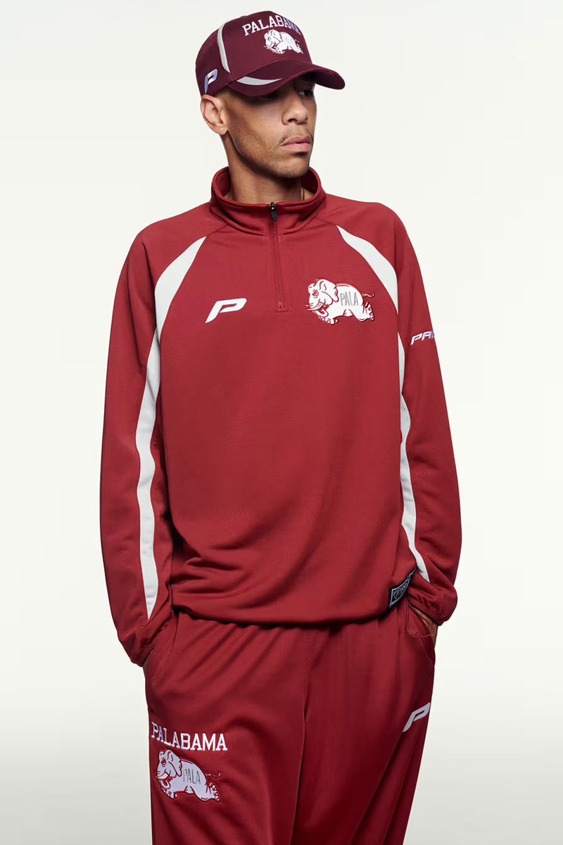 Man in matching maroon sweat suit with white logo and hat poses in photo studio, showcasing casual sportswear apparel.