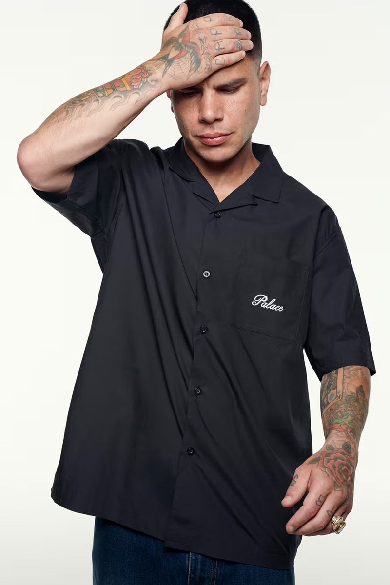 Man with tattoos posing in a black Palace button-down shirt against a white backdrop, indoor studio setting, looking away from camera.