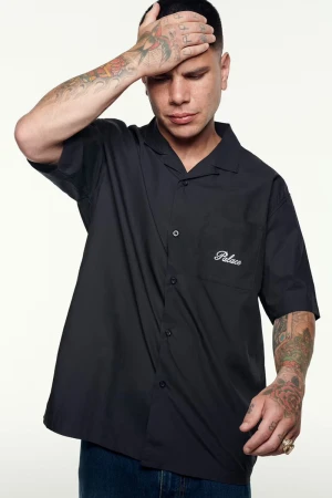 Man with tattoos posing in a black Palace button-down shirt against a white backdrop, indoor studio setting, looking away from camera.