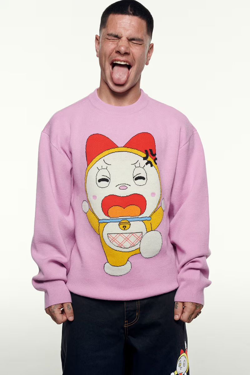 Man with his tongue out wears a pink sweater with a cartoon character on it. He has short dark hair and is wearing dark jeans.