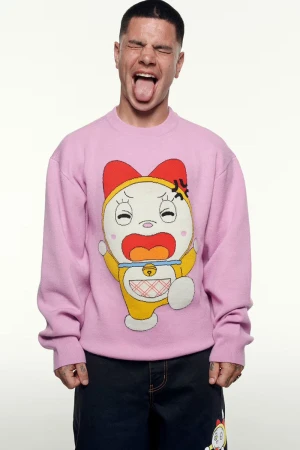 Man with his tongue out wears a pink sweater with a cartoon character on it. He has short dark hair and is wearing dark jeans.