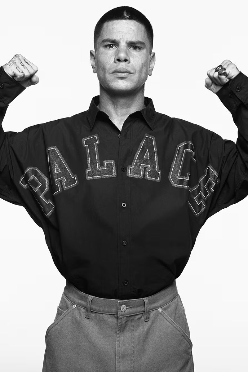 A person in a black Palace brand shirt flexes their arms in a studio shot. The individual wears denim jeans and sports a unique crescent tattoo.