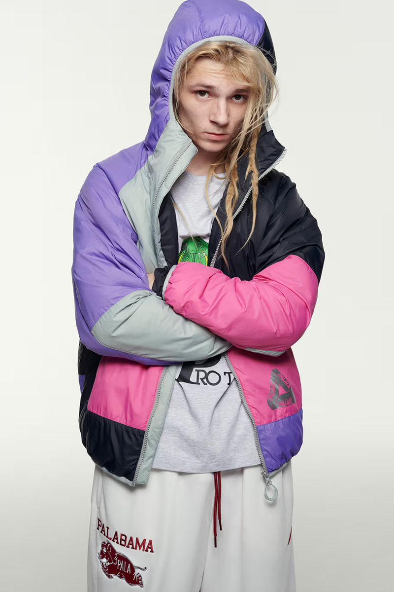 Young adult with long blonde hair poses wearing a color-blocked puffer jacket and white sweatpants in a brightly lit photo studio.