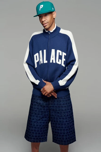 A man in blue Palace brand zip-up sweater with white accents and blue patterned shorts poses against a grey backdrop in a studio setting.