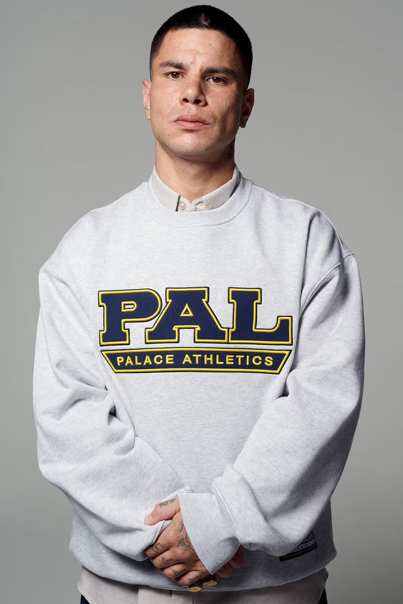 Portrait of a man with short dark hair wearing a grey Palace Athletics sweatshirt, posing in front of a neutral grey background.
