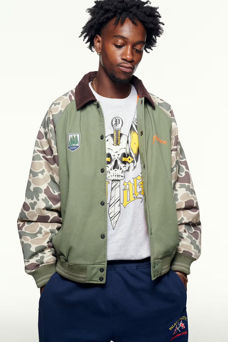 A young adult Black male poses indoors, wearing an olive green jacket with camouflage sleeves and blue sweatpants, against a neutral backdrop.
