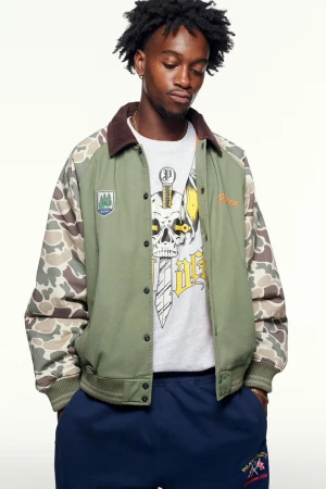 A young adult Black male poses indoors, wearing an olive green jacket with camouflage sleeves and blue sweatpants, against a neutral backdrop.