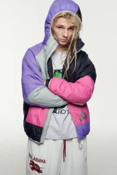 Young man with blonde hair wearing a color-blocked puffer jacket and white trousers against a plain white background