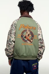 Man wearing a green camouflage-sleeved jacket with Palace branding on the back against a white seamless background.