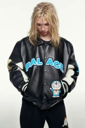 Model with long blonde hair wearing a black leather jacket with blue patch lettering against a white seamless backdrop.