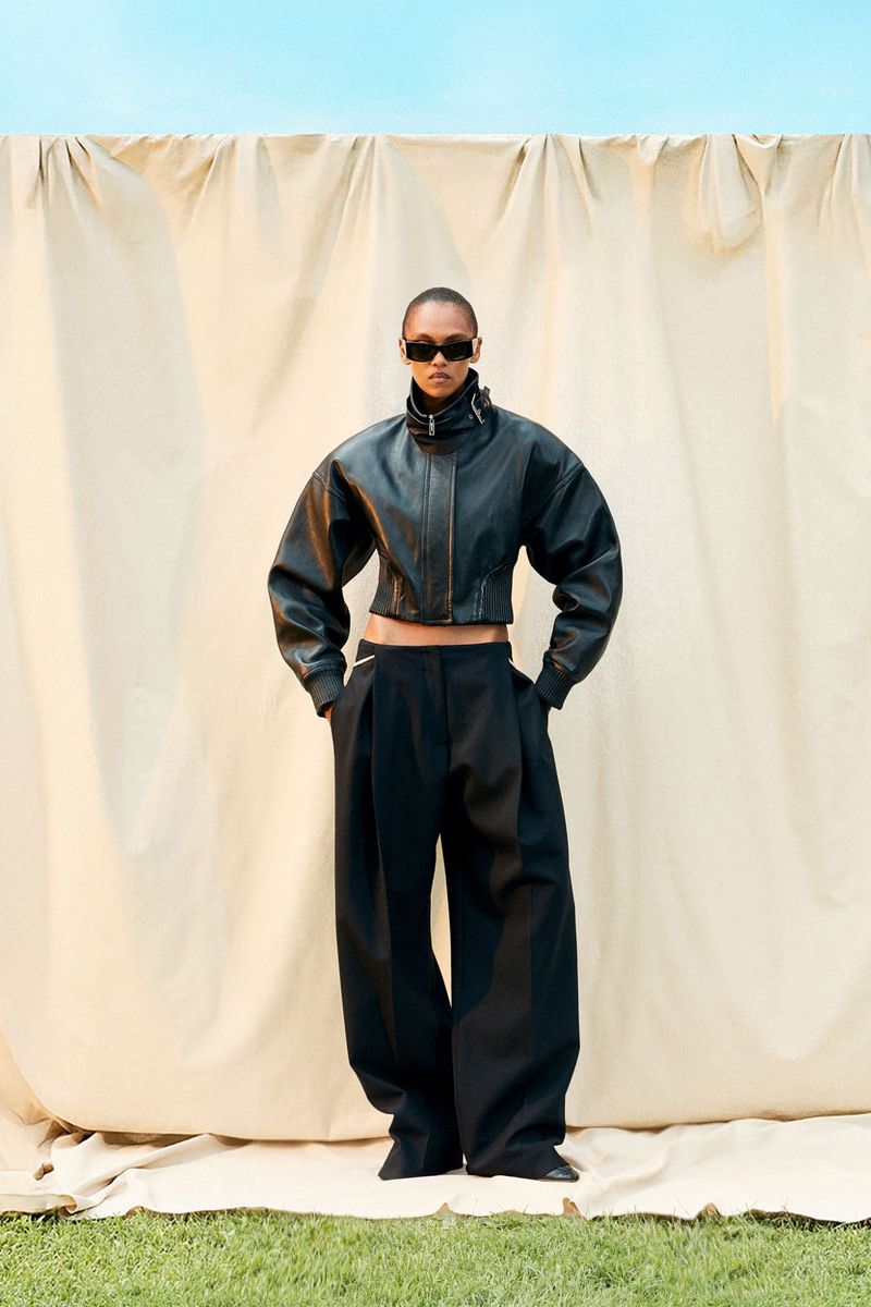 A fashionable person in a black leather jacket and wide-leg pants stands outdoors against a tan backdrop on a grassy surface.
