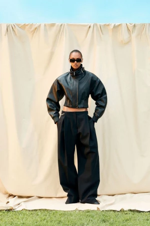 A fashionable person in a black leather jacket and wide-leg pants stands outdoors against a tan backdrop on a grassy surface.