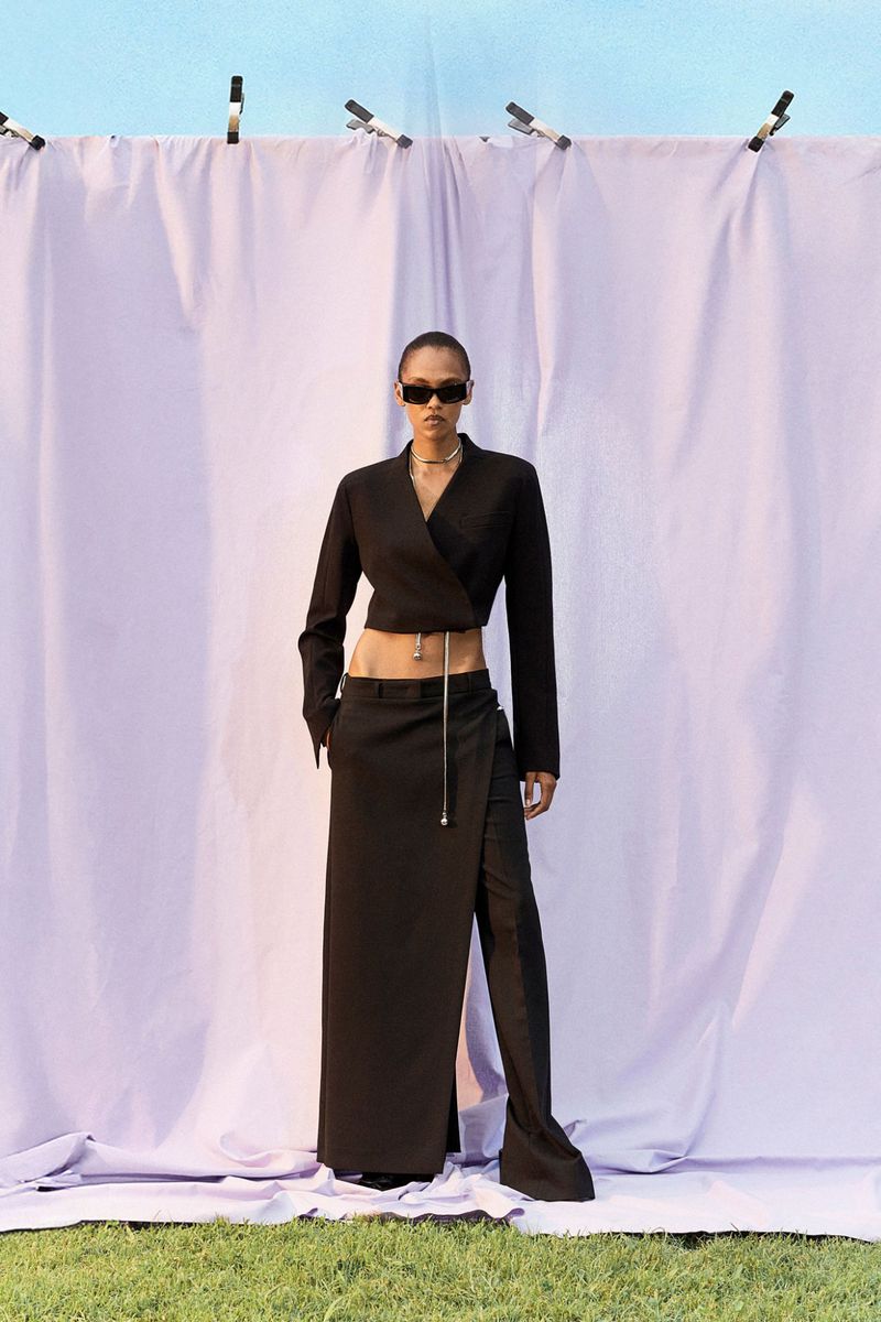 Adult female fashion model in an outdoor setting. She wears a black, short jacket, long skirt, and sunglasses, with a lavender backdrop.