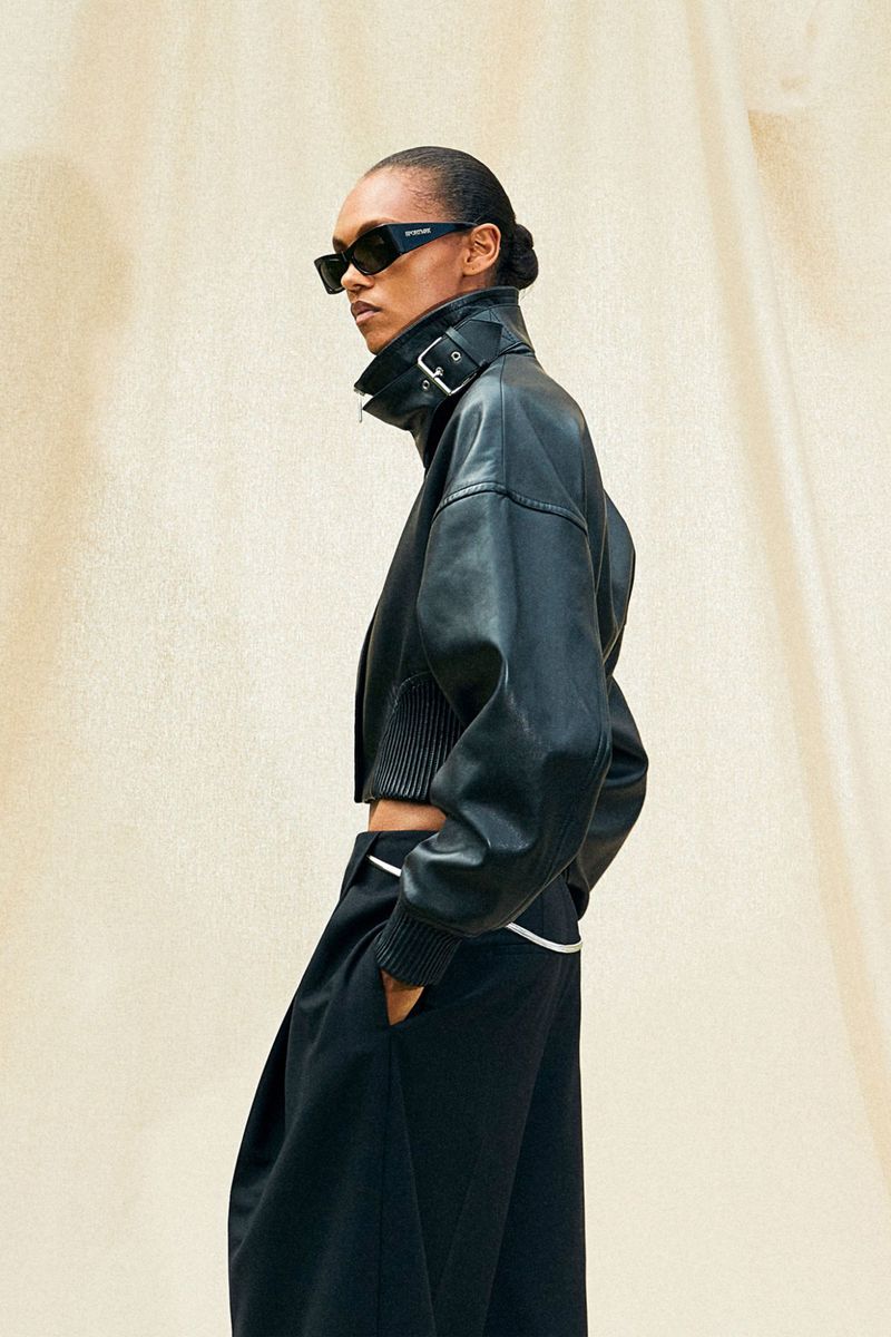 Adult fashion model poses in a black leather jacket, black sunglasses, and black skirt against an off-white neutral studio backdrop.