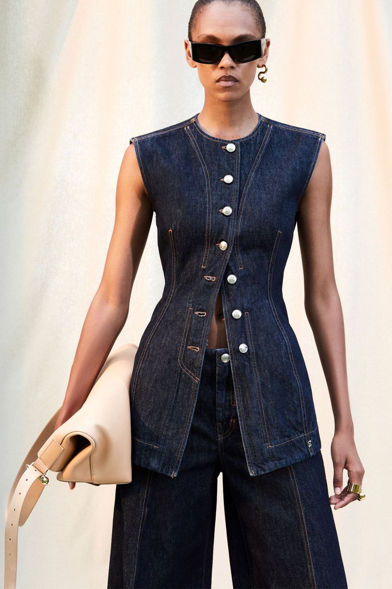Woman in denim vest and culottes holding a tan clutch bag, wearing sunglasses and jewelry, posed against a neutral backdrop.