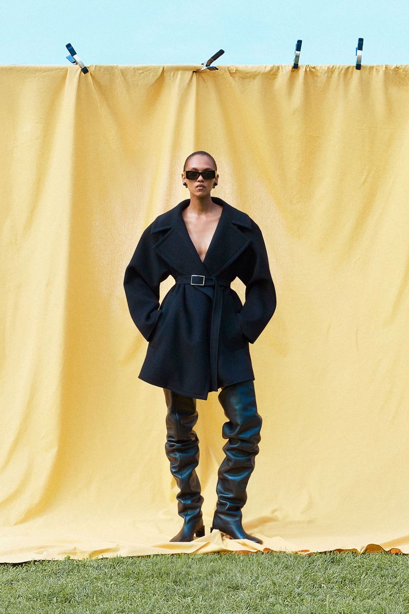 Fashion editorial shot of a woman in a black coat and thigh-high boots posing in front of a yellow curtain, with an outdoor background.
