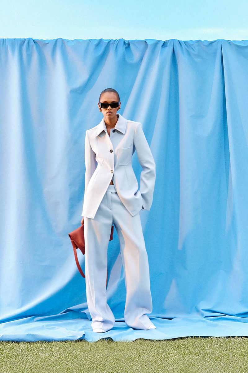 A woman in a light blue pantsuit stands in front of a blue sheet holding a brown purse, creating a chic and modern fashion moment.