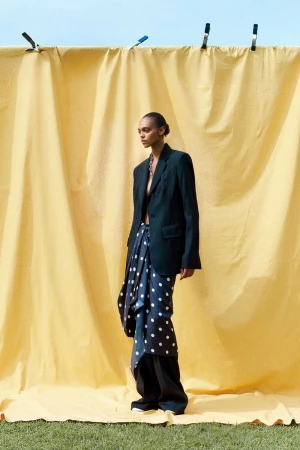 Fashion shot of a slender woman in a blazer and polka-dot skirt standing in front of a yellow fabric backdrop on green grass.